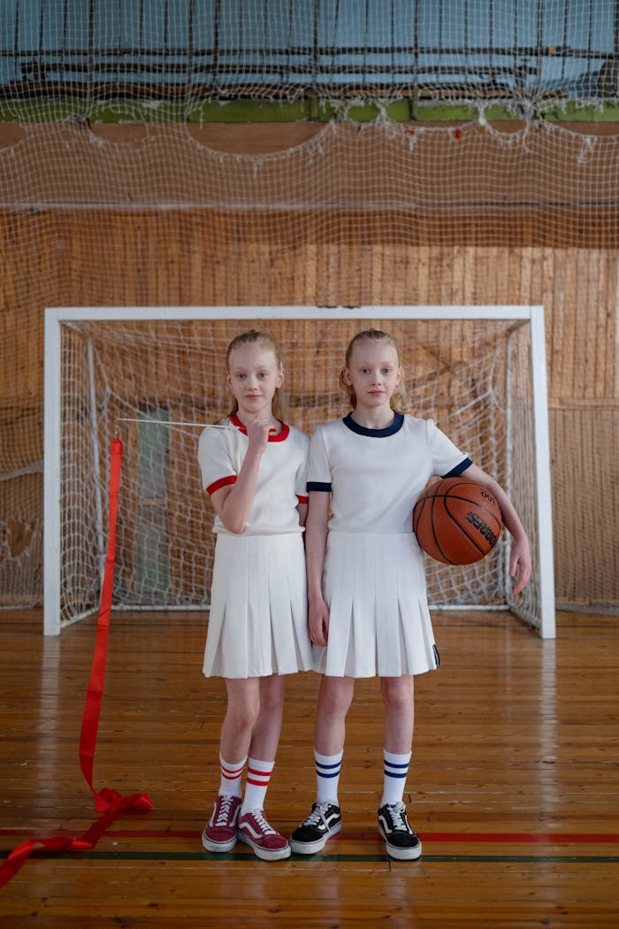 Twin girls in matching sports uniforms standing with a basketball indoors.